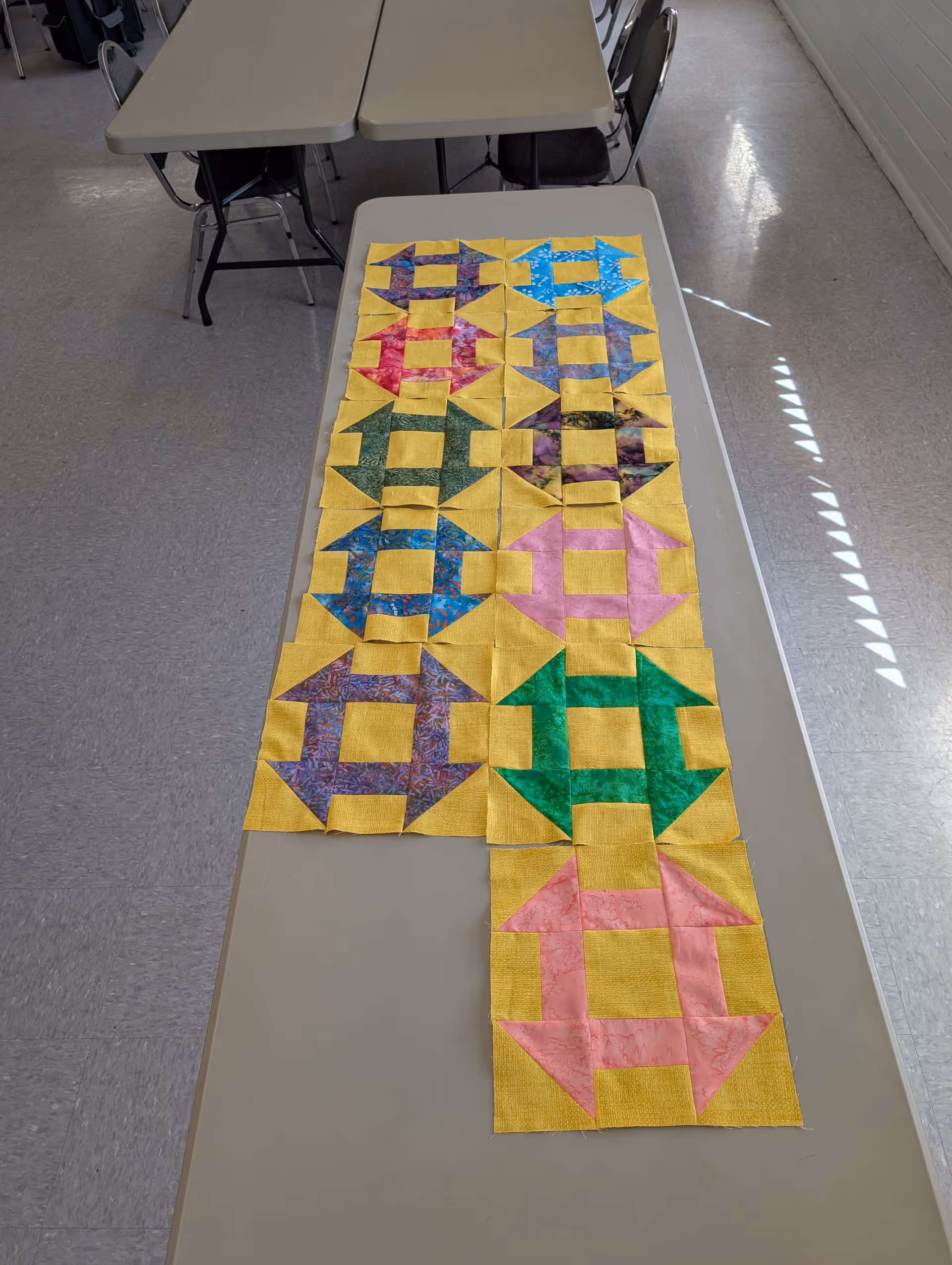 Colorful quilt layout on a long table in a bright indoor room.