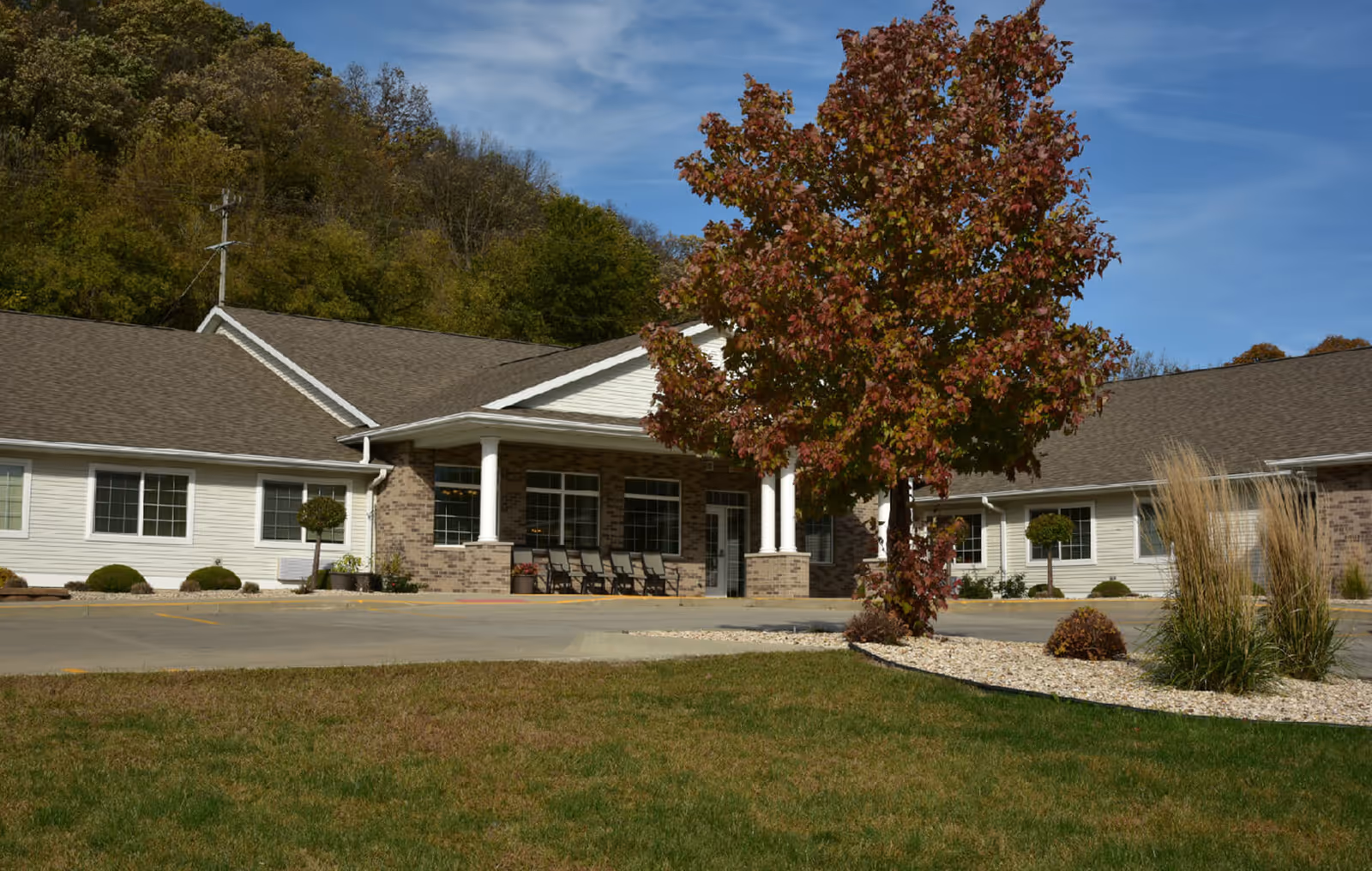 Exterior view of Kingston Court Assisted Living facility showing a single-story building with a covered entrance supported by white columns. There is a tree with autumn-colored leaves and landscaped bushes in front of the building under a partly cloudy sky.