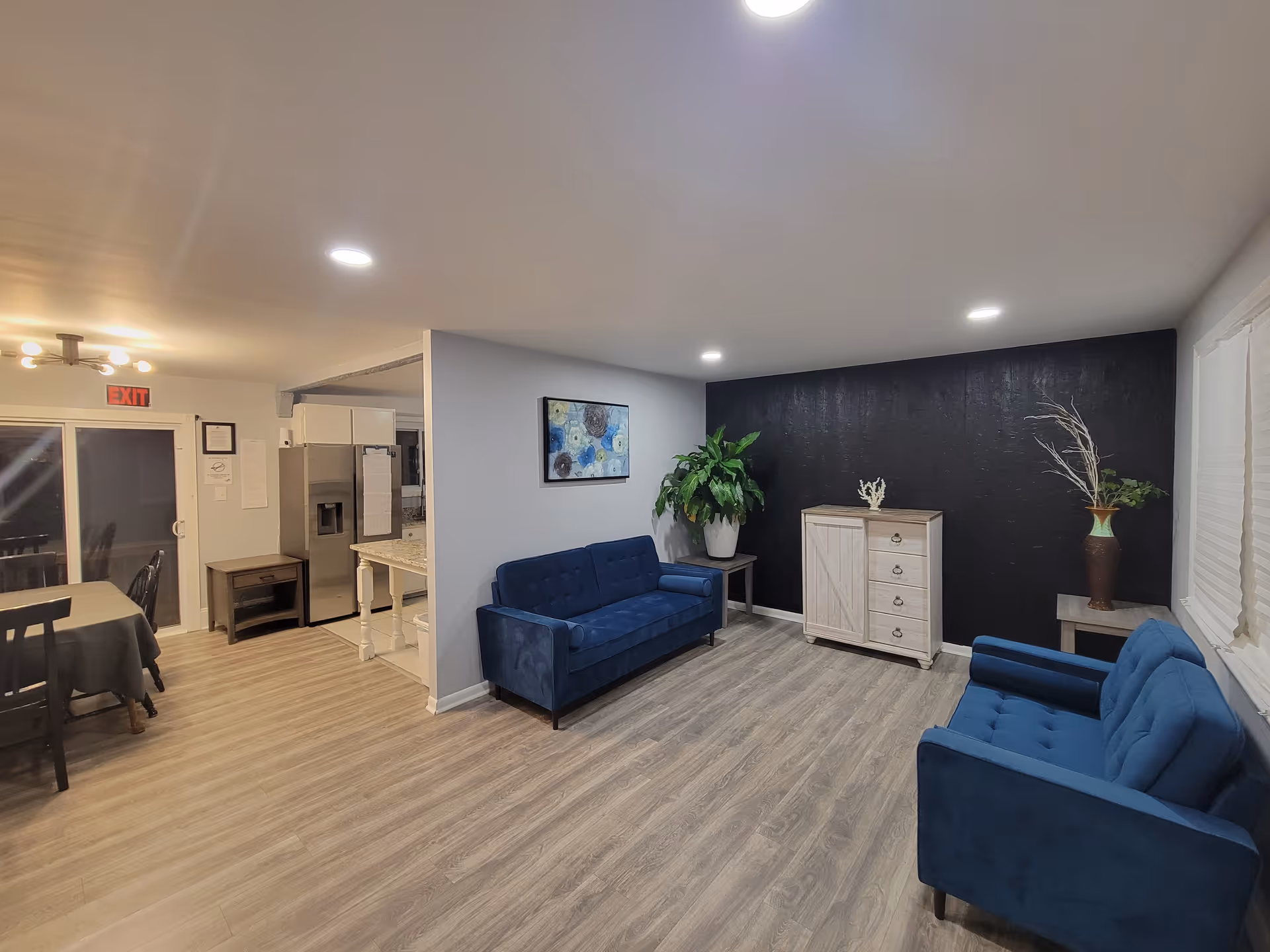 Bright lounge area with blue sofas, plants, and an open dining-kitchen doorway.