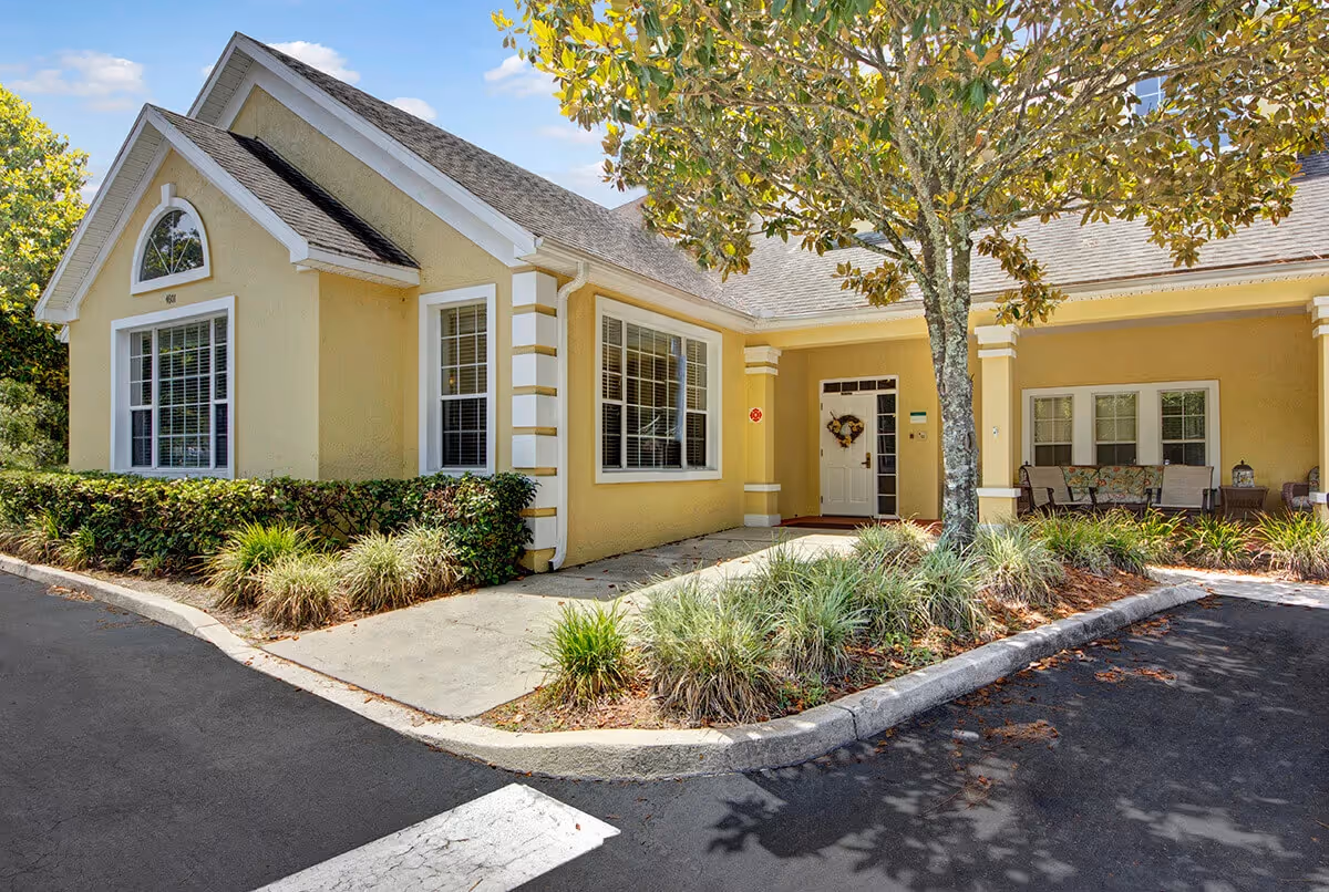 Front entrance of a yellow single-story building with a covered porch, landscaping, and a tree in the driveway.