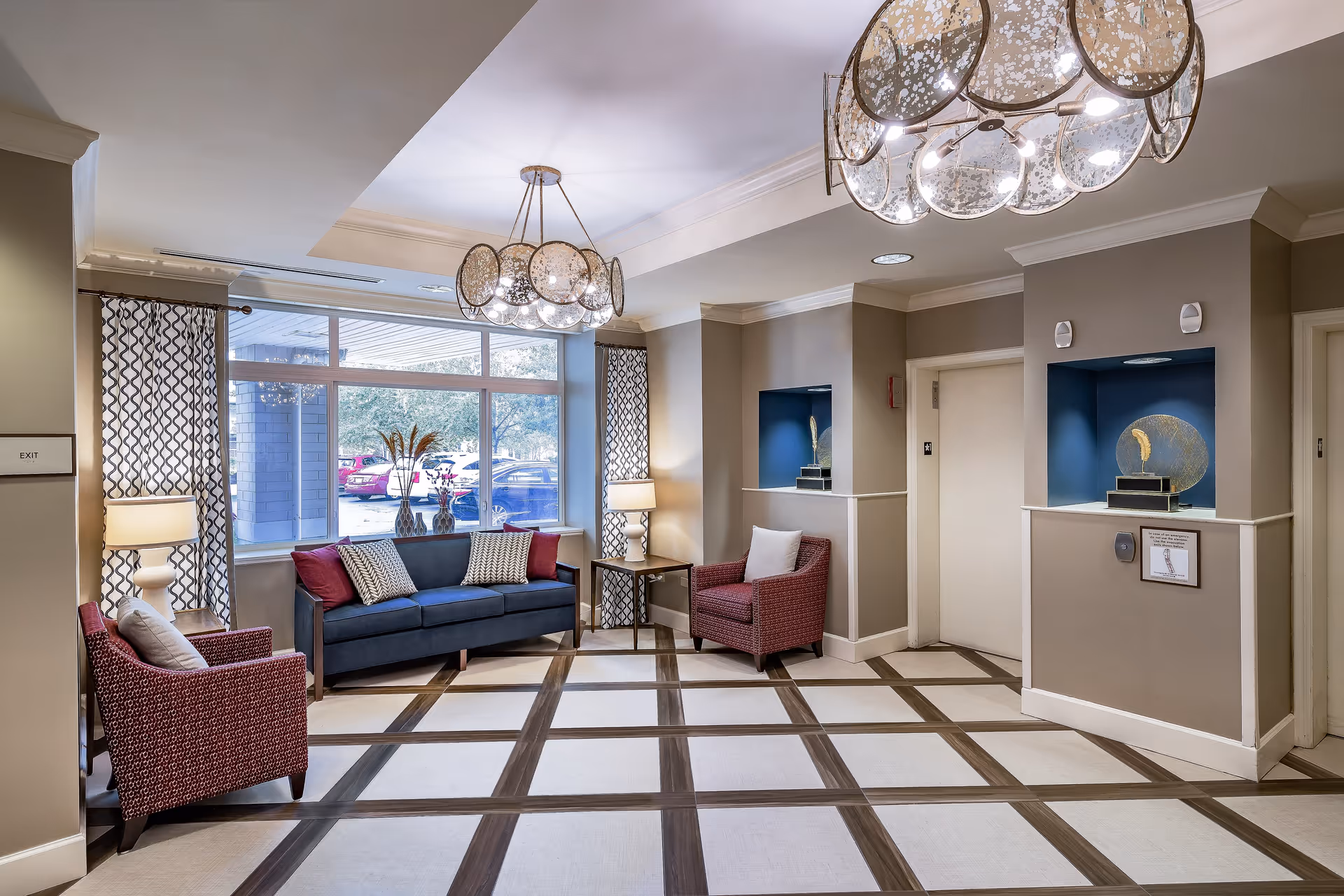 A well-lit seating area in a senior living facility with a blue sofa and two red armchairs arranged around a small wooden table. The room features large windows with patterned curtains, two modern lamps, decorative wall niches with art pieces, and a stylish chandelier hanging from the ceiling. The floor has a checkered pattern with light and dark tiles.