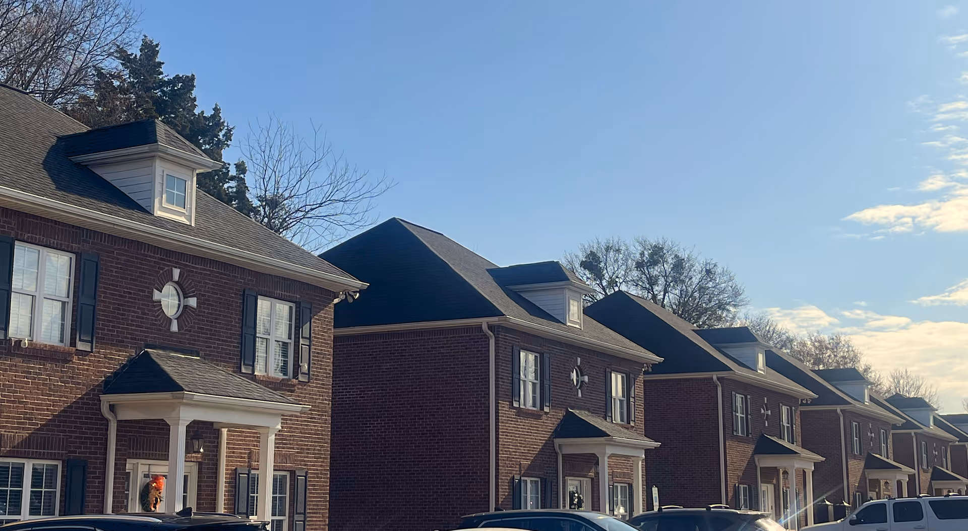 Row of brick townhouses with dark roofs and parked cars along the street.
