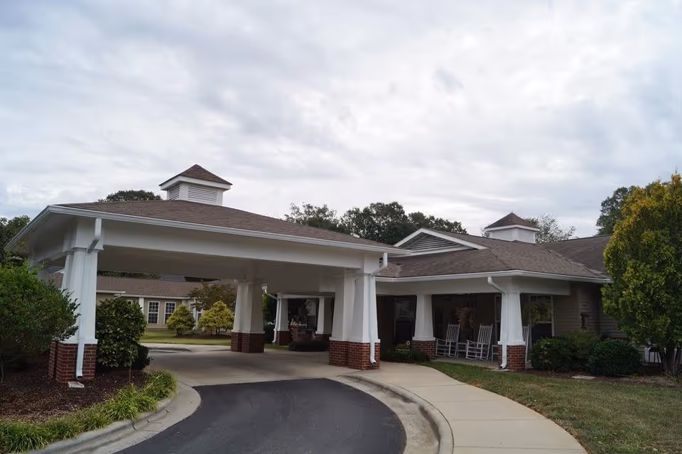 Exterior view of Somerset Court of Cherryville showing a covered driveway entrance with white columns and brick bases. The building has a brown shingled roof with cupolas and is surrounded by greenery including bushes and trees. There is a curved driveway and a sidewalk leading to the entrance.