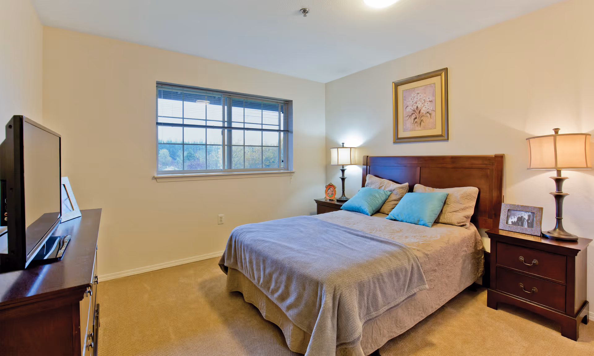 Bedroom in a senior living facility with bed, nightstands, lamps, and TV.