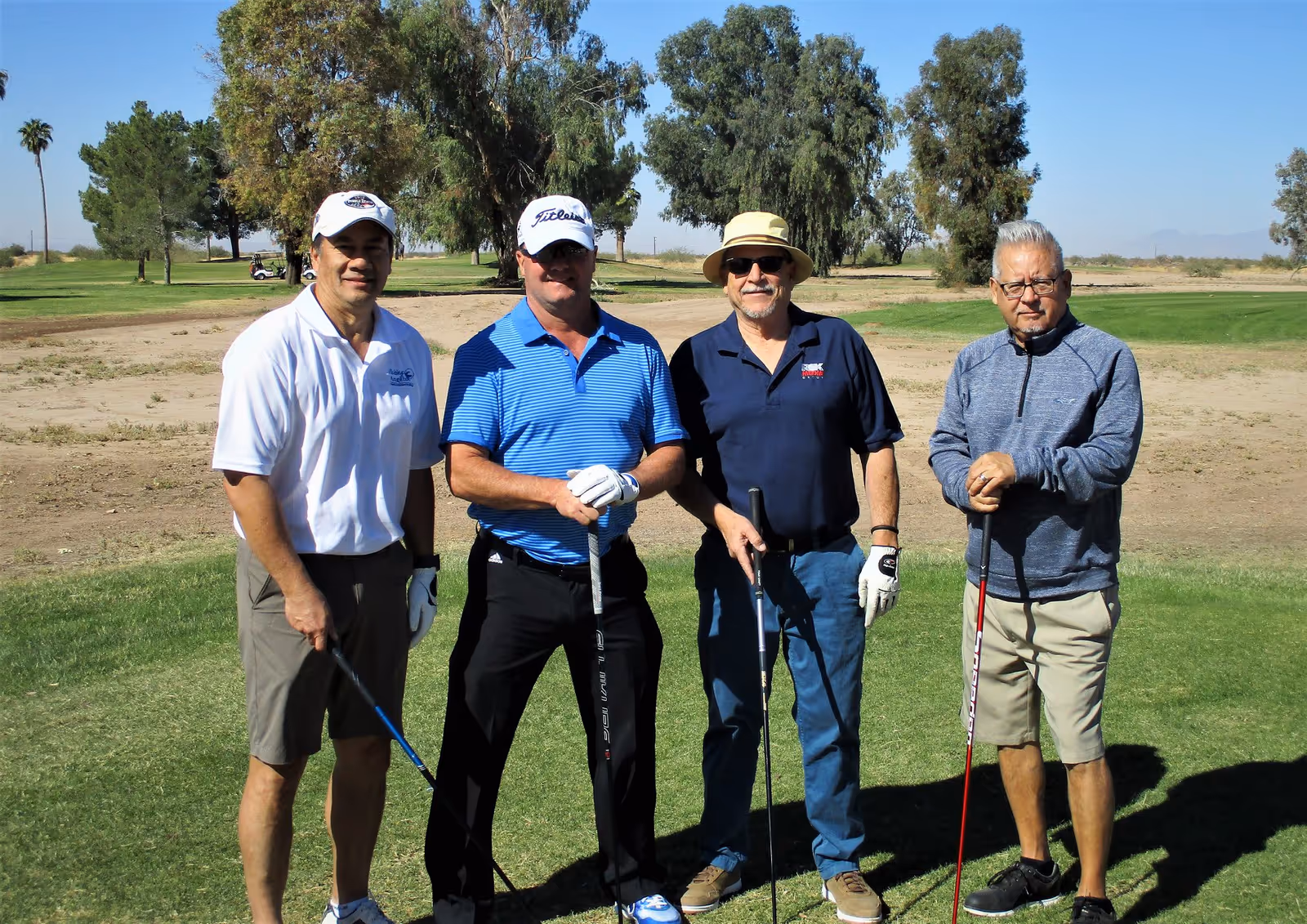 Four men in golf attire pose with clubs on a sunny outdoor golf course