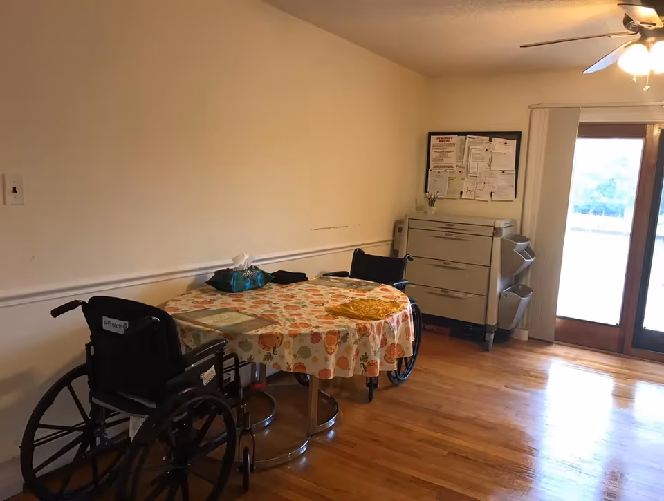 A small room with a round table covered with a floral tablecloth, two wheelchairs positioned around the table, a tissue box on the table, a filing cabinet with papers pinned on a bulletin board above it, and a glass door letting in natural light.