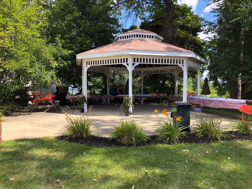 White gazebo with red-check tables under trees in a sunny outdoor gathering area.