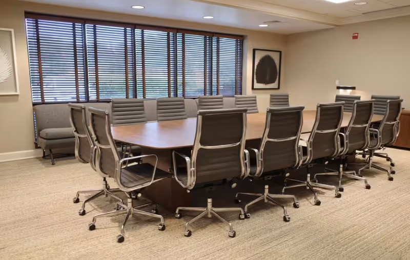 A modern conference room with an oval table, numerous rolling chairs, and window blinds.