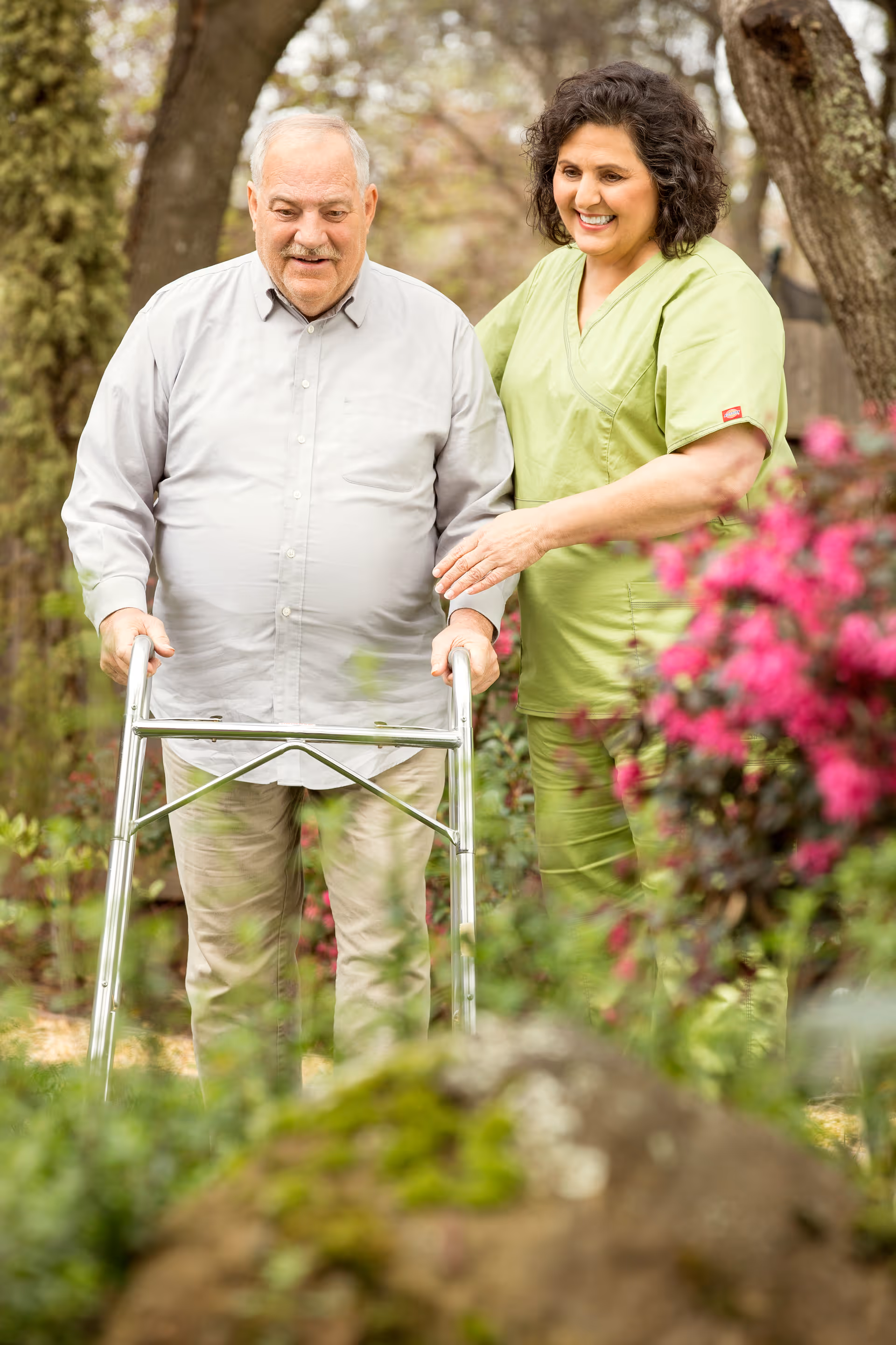 An elderly man with a walker is guided by a caregiver through a garden path.