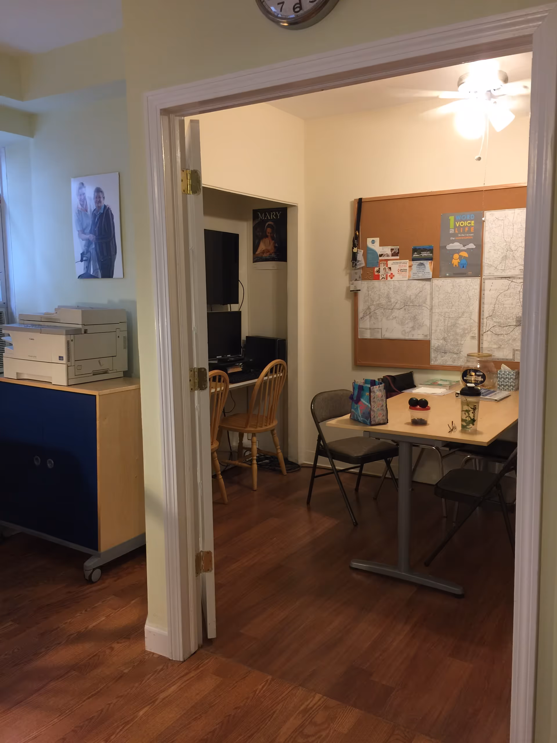 Interior room with bulletin board, table, chairs, and computer area beyond doorway.