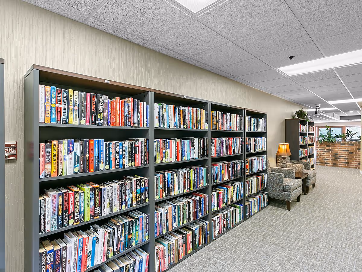 Interior view of a library or reading area in a senior living facility with multiple bookshelves filled with books. There are two patterned armchairs with a small table and lamp between them. The space has carpeted flooring, a drop ceiling with fluorescent lights, and a brick half-wall with plants in the background.
