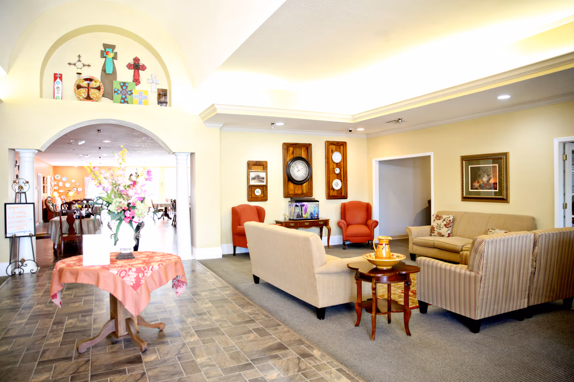 Bright, well-lit common sitting area with sofas and armchairs, a table with flowers in the foreground, and an arched opening to a dining area.