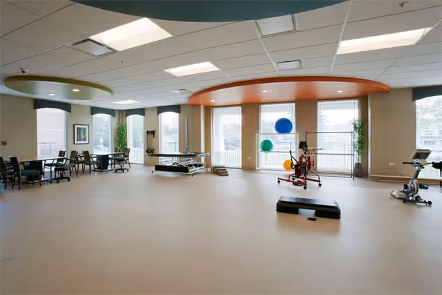 Spacious therapy/exercise room with treatment tables, exercise equipment, stability balls, and seating under circular ceiling accents and large windows.