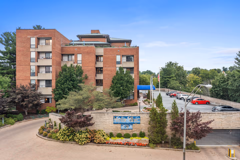 Exterior view of Mayfair Village Retirement Community, a multi-story brick building surrounded by trees and landscaping. There is a parking area with several cars and a sign at the entrance displaying the facility's name and address.