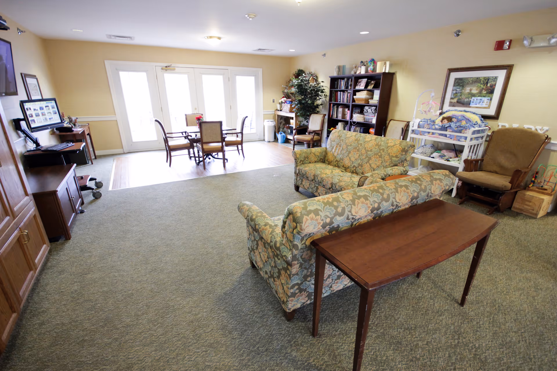 A spacious senior living lounge with seating, tables, a bookshelf, and a crib.
