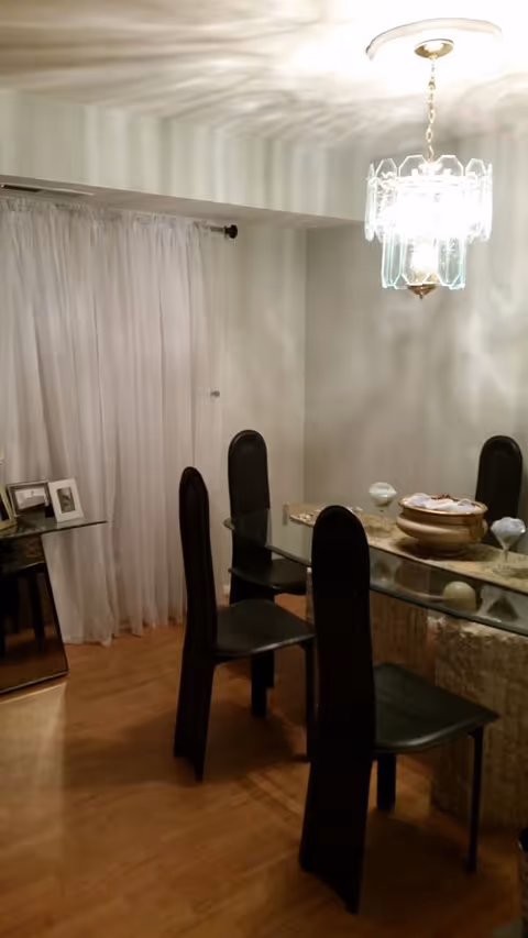 A glass dining table with black chairs sits under a chandelier in a bright dining room.