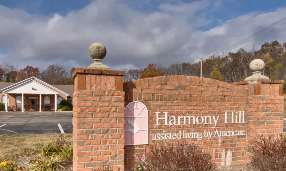 Exterior view of Harmony Hill assisted living facility with a brick sign in the foreground displaying the facility's name and a building with a covered entrance in the background under a cloudy sky.