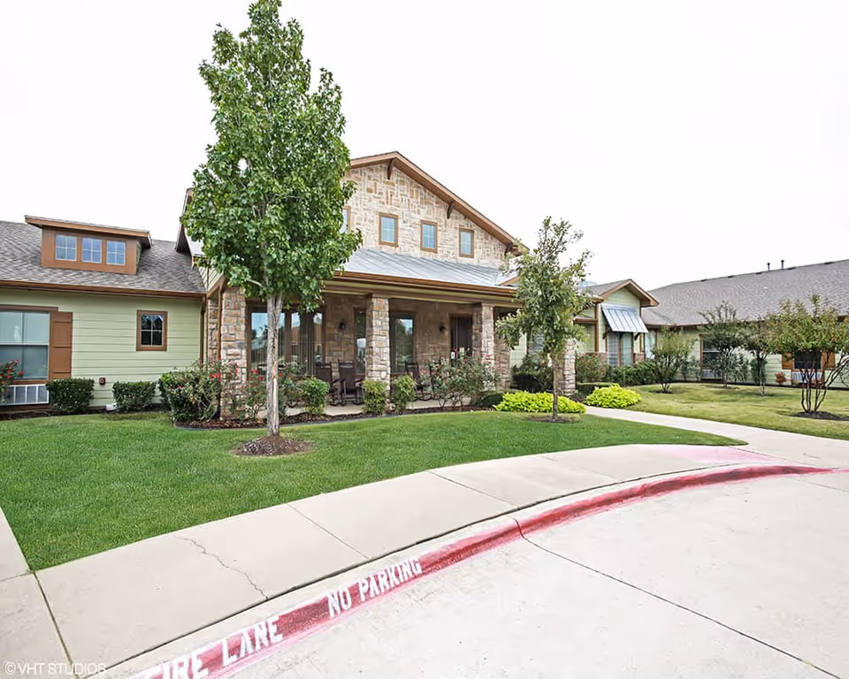 Front entrance of a single-story assisted living building with a stone porch, landscaping, and a curved driveway.