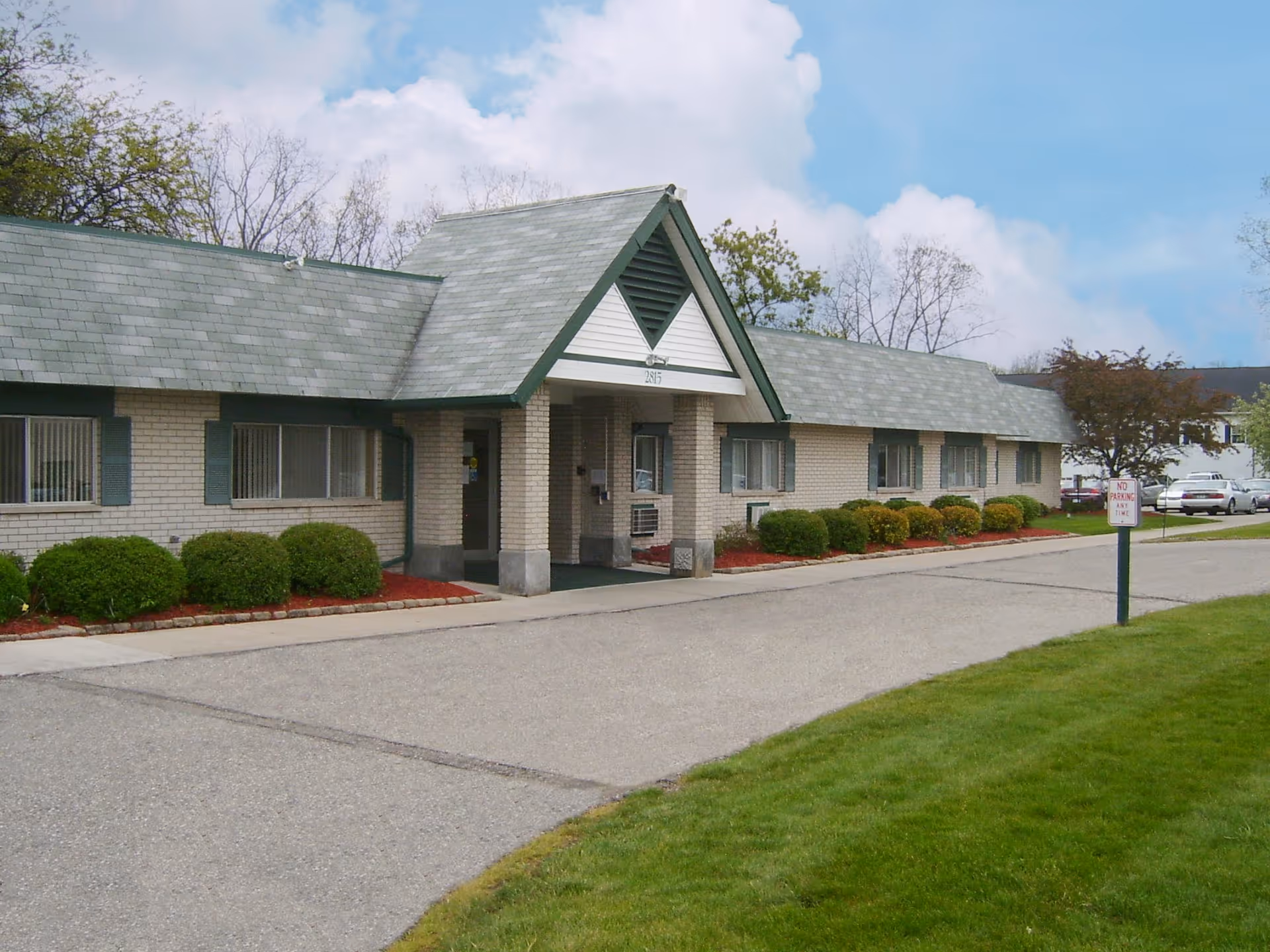 Exterior view of a single-story brick building with a green shingled roof and a covered entrance. The building is surrounded by neatly trimmed bushes and a paved driveway. There is a 'No Parking Any Time' sign on a post near the driveway, and several cars are parked in the background. The sky is partly cloudy.