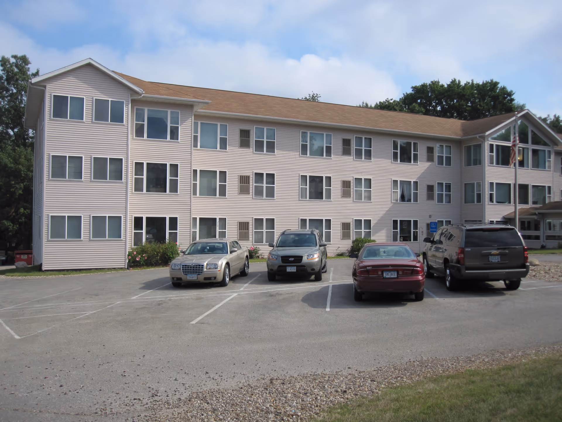 Front exterior of a three-story beige apartment-style senior living building with several cars parked in the lot.