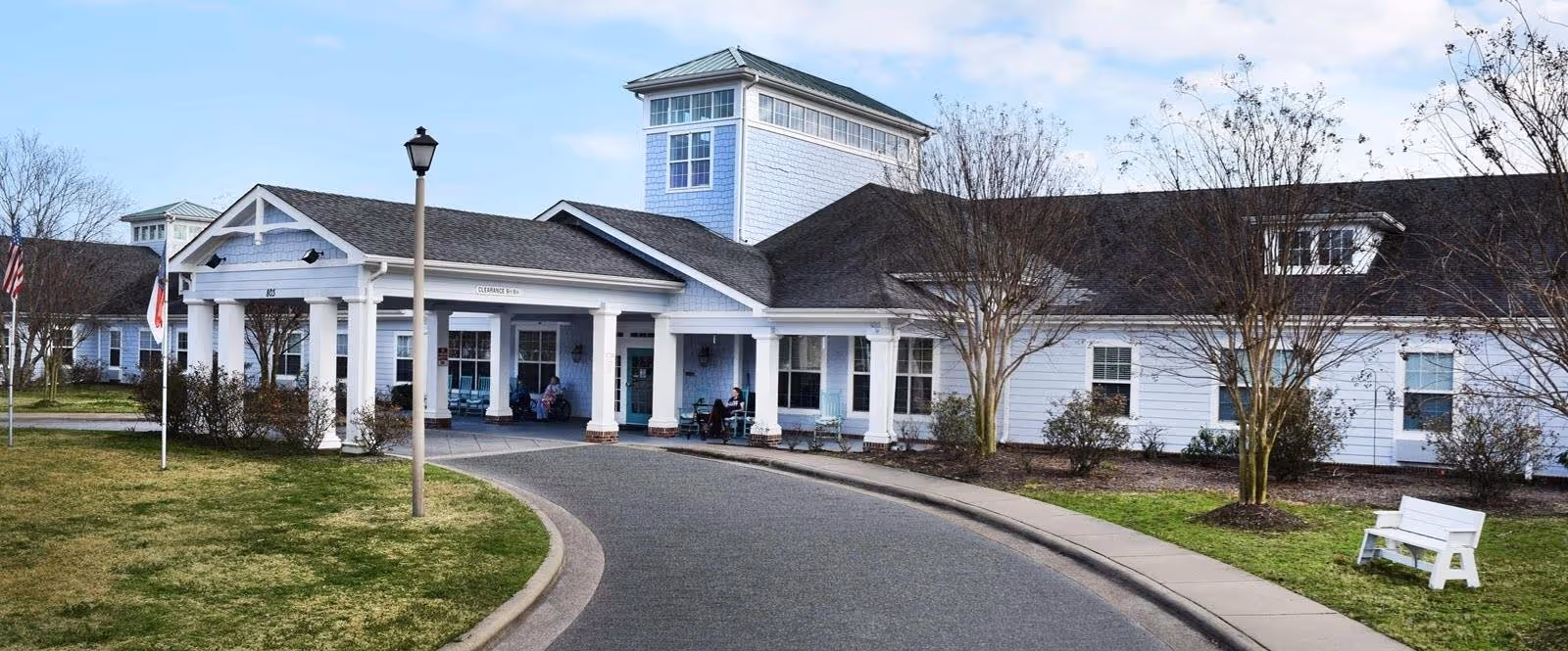 The front entrance of a white senior living facility with a covered portico, curved driveway, lawn, and benches.