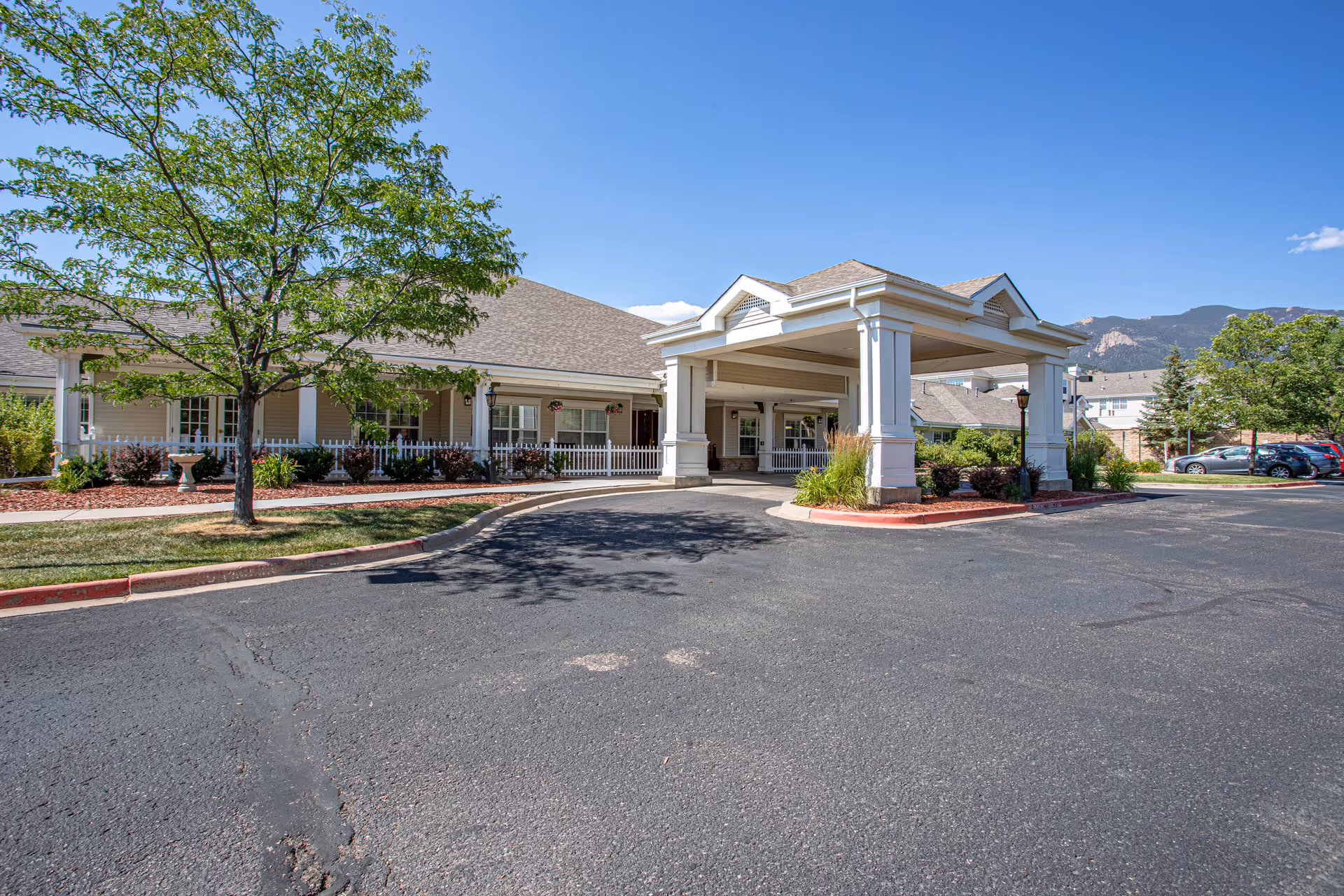 Exterior view of Bear Creek Assisted & Senior Living facility showing the main entrance with a covered drop-off area, surrounded by landscaped greenery and a parking lot under a clear blue sky.