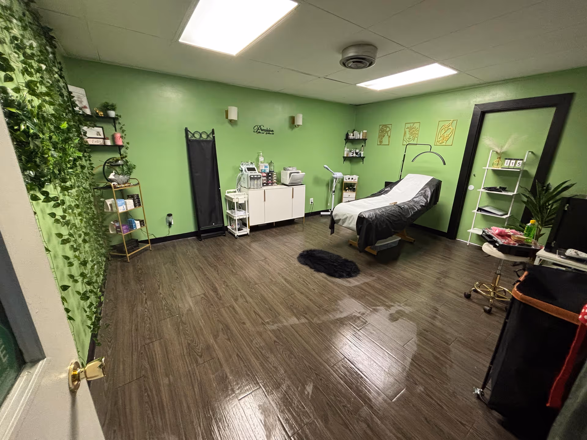 A green treatment room with a reclining bed, medical carts, shelves, and plants.