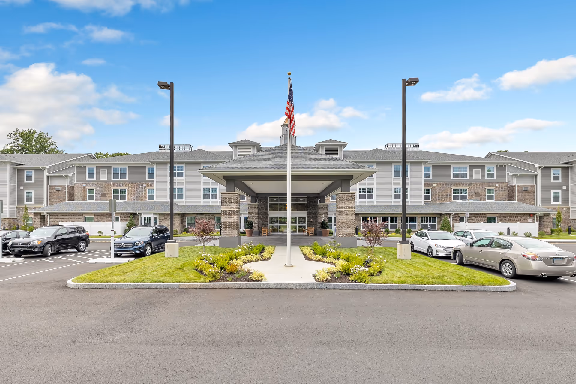 Front entrance of a senior living community building with a covered drop-off, American flag, parked cars, and landscaped driveway.