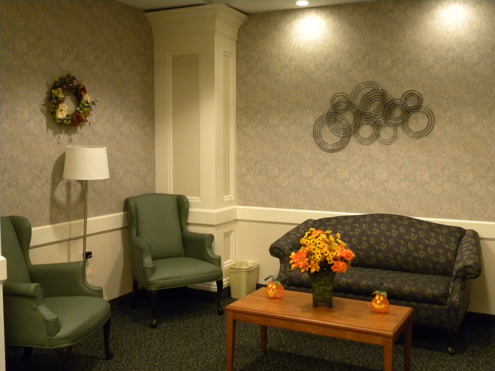 A cozy sitting area with two green upholstered armchairs and a dark patterned sofa. A wooden coffee table in front of the sofa holds a vase with orange and yellow flowers and two small pumpkin decorations. The walls are decorated with a floral wreath and a modern metal wall art piece. A floor lamp stands beside one of the armchairs.