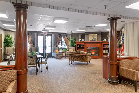 Spacious senior living lounge with couches, chairs, fireplace, bookshelves, and decorative columns.