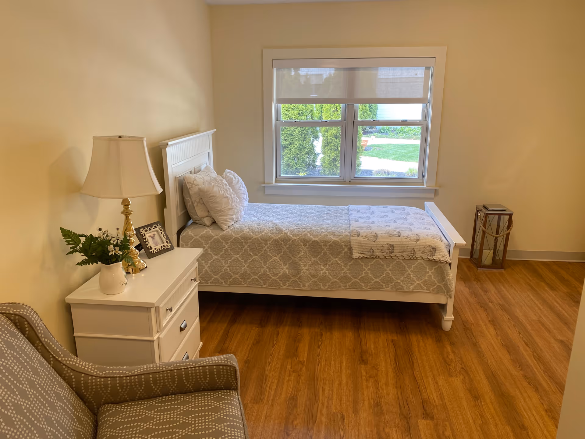 This bright Seasons of Danvers bedroom features a white bed, nightstand, lamp, chair, and window view.