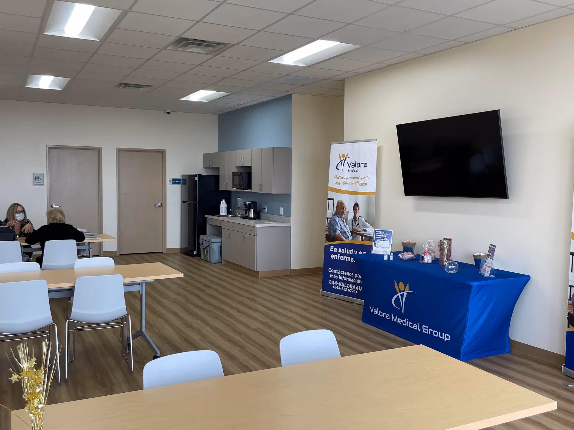 Bright facility room with kitchenette, tables, chairs, two masked people, and a Valora banner.