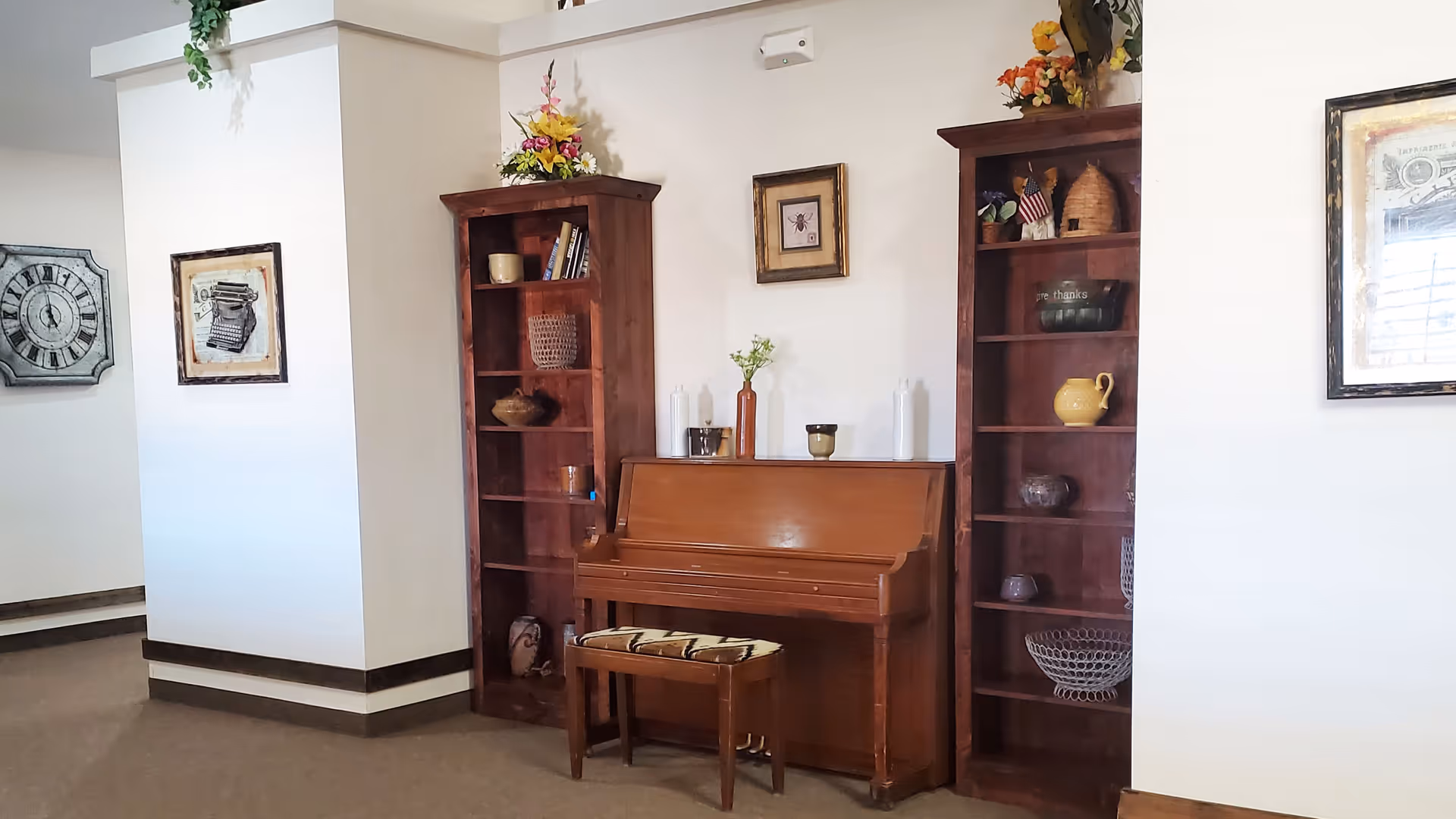 A cozy interior corner featuring a wooden upright piano with a cushioned bench in front. On either side of the piano are tall wooden bookshelves filled with decorative items such as vases, baskets, and small flags. Above the piano hangs a small framed picture, and there are additional framed artworks on the adjacent walls. The room has light-colored walls and carpeted flooring.