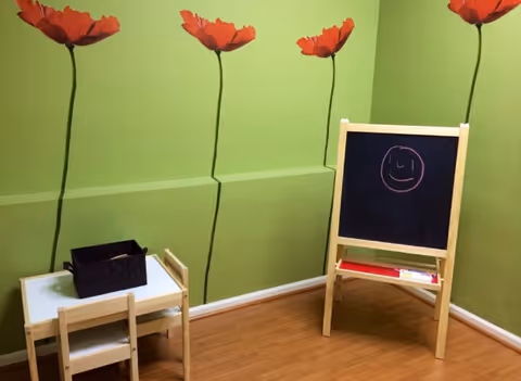 Small play corner with green walls, flower decals, chalkboard easel, and kid-sized furniture.