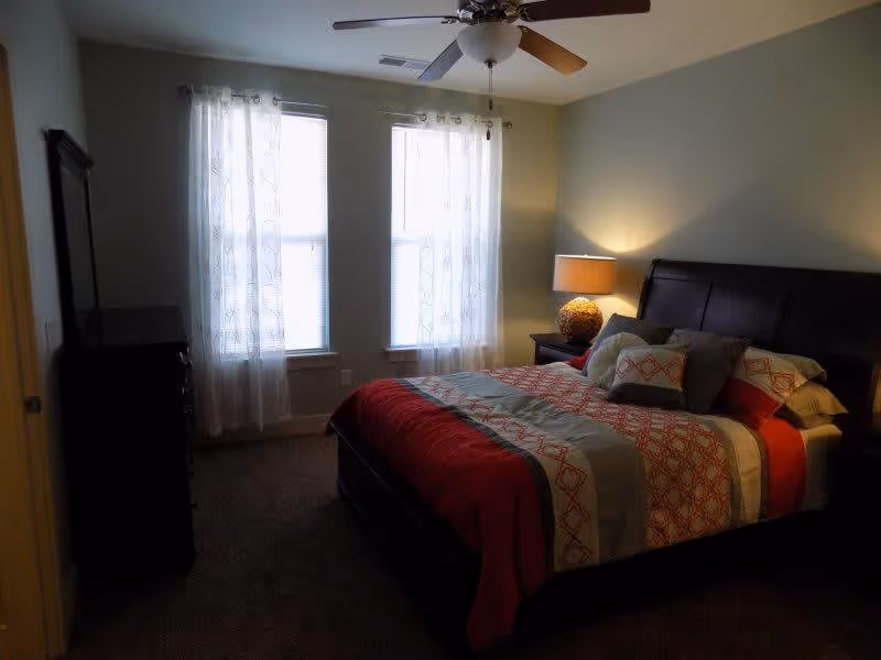 A bedroom with a large bed featuring a red and white patterned comforter and multiple pillows. There is a nightstand with a lamp on the right side of the bed, two windows with sheer white curtains letting in natural light, a ceiling fan above, and a dark wooden dresser on the left side of the room.