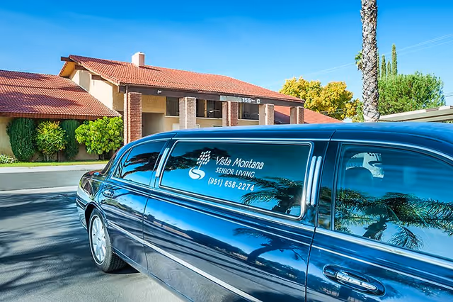 A black limousine bearing a 'Vista Montana Senior Living' logo is parked in front of a single-story senior living building.