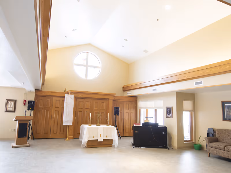 A bright room with a high ceiling featuring a round window. There is a small altar covered with a white cloth and three candles in the center. To the left, there is a wooden podium and two speakers. On the right side, there is a black piano and a beige couch with a green plant nearby. The walls are light-colored with wooden trim and some framed pictures.
