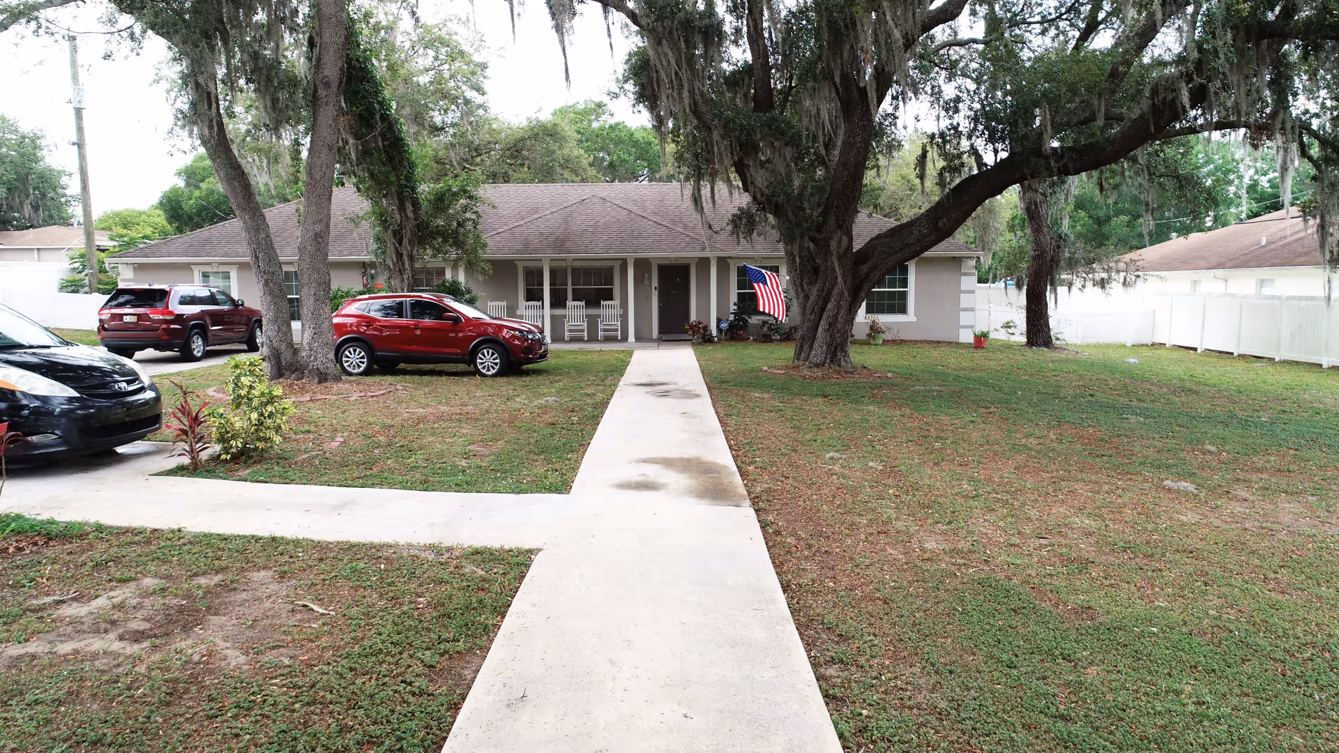Front exterior view of a single-story residential building with a covered porch featuring four white rocking chairs. There are three cars parked on the driveway and lawn, large trees with hanging moss, an American flag near the entrance, and a concrete walkway leading to the front door. The yard is grassy with some small plants and a white fence in the background.
