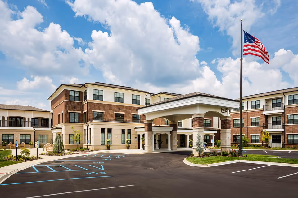 Exterior view of a three-story senior living facility with a covered entrance, brick and beige facade, multiple windows, an American flag on a flagpole, and a parking lot with marked spaces including handicapped spots.