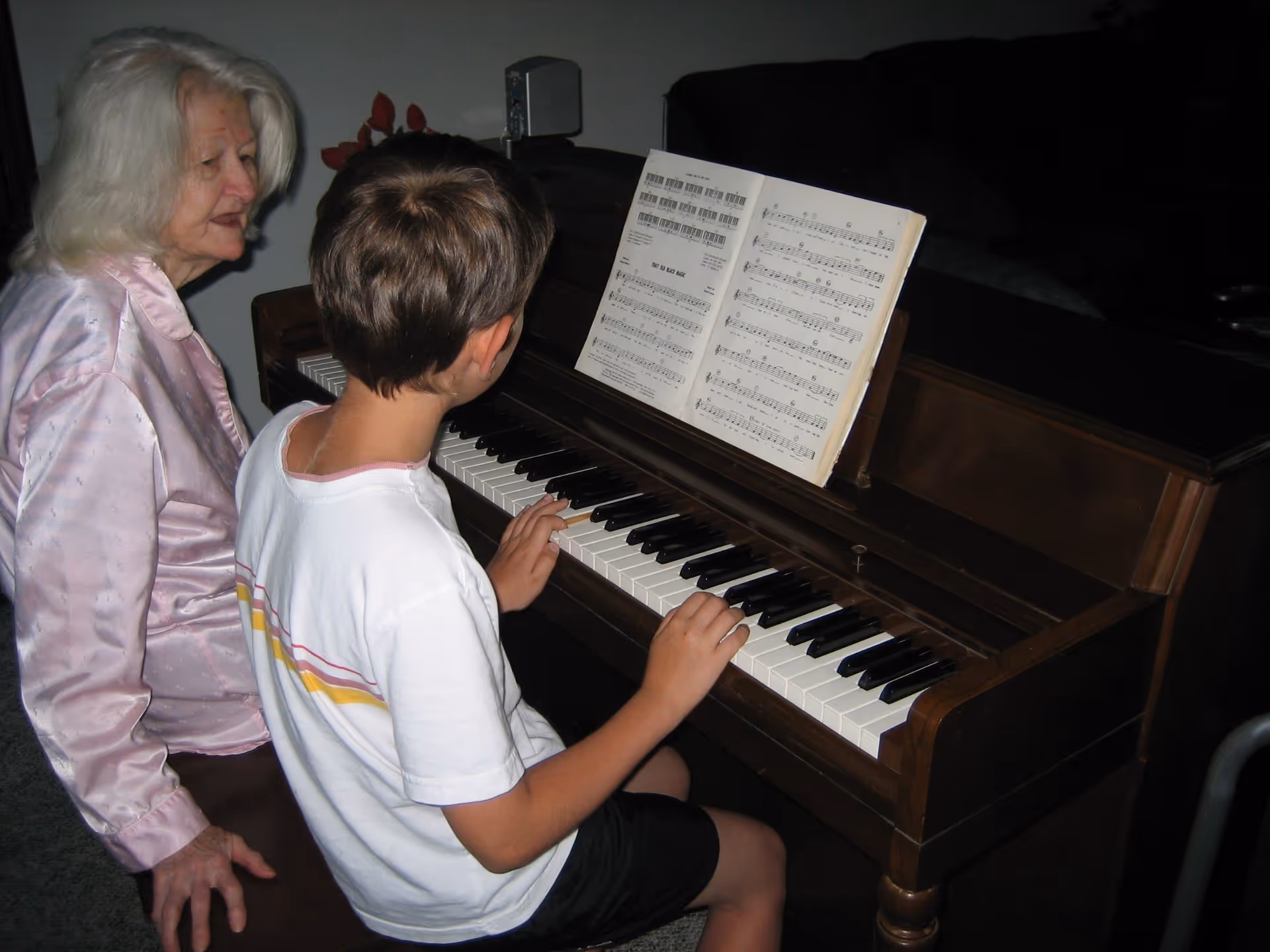 An elderly woman with white hair and a pink satin blouse sits beside a young boy playing a wooden piano. The boy is reading sheet music placed on the piano stand. They are in a dimly lit room with a dark couch in the background.