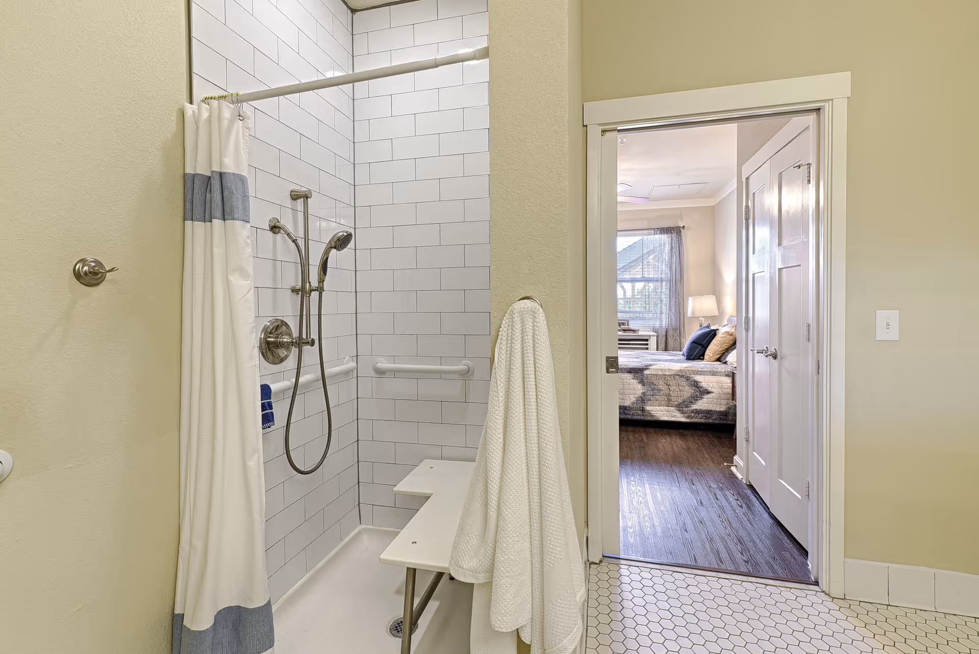 Accessible bathroom with tiled shower, bench, grab bars, and doorway to a bedroom.