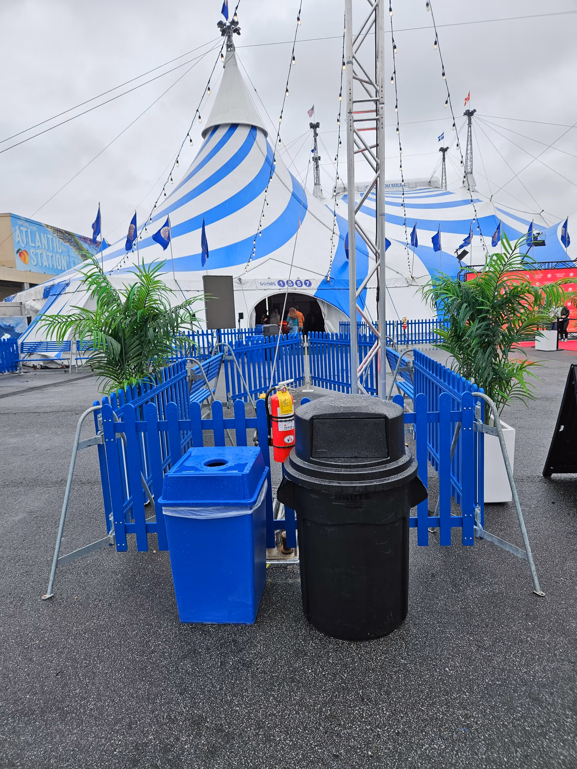 Blue-and-white striped circus tents with blue fencing, trash bins, and potted plants outdoors.