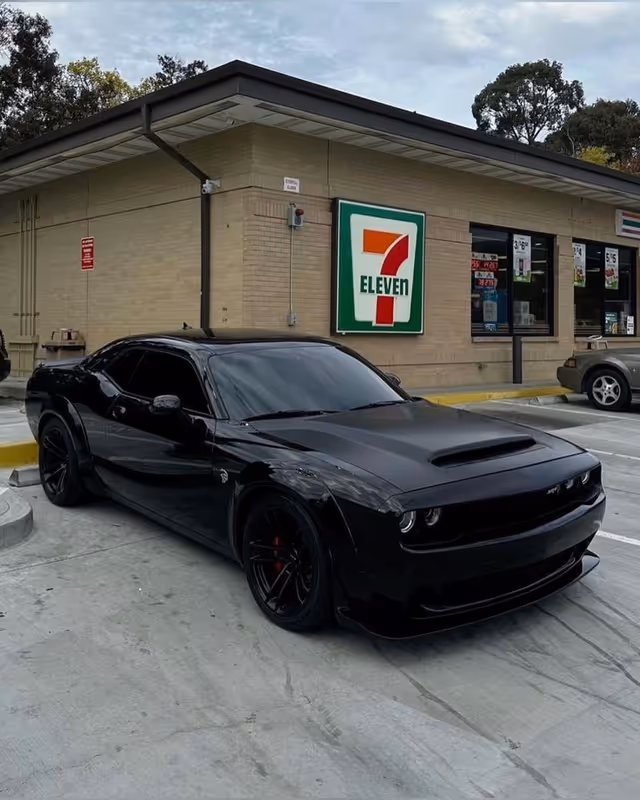 A black sports car parked in a lot beside a beige building with a 7-Eleven sign.