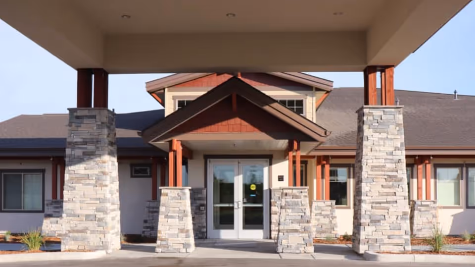 Front entrance of a senior living facility with stone pillars and a covered driveway. The building has a peaked roof and large glass double doors.