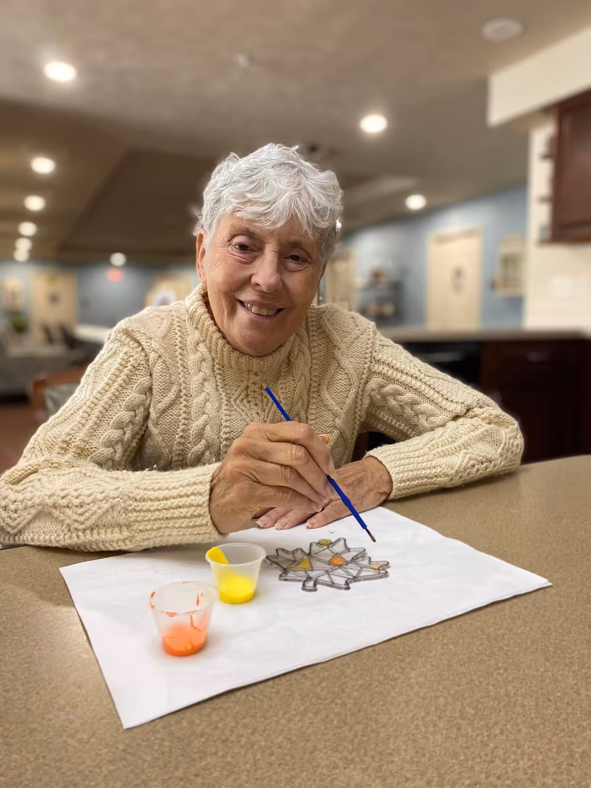 An elderly woman with short white hair wearing a cream-colored cable knit sweater is sitting at a table, smiling and painting a stained glass design on a piece of paper. There are small cups of yellow and orange paint on the table. The background shows a softly lit interior space with blurred furniture and walls.
