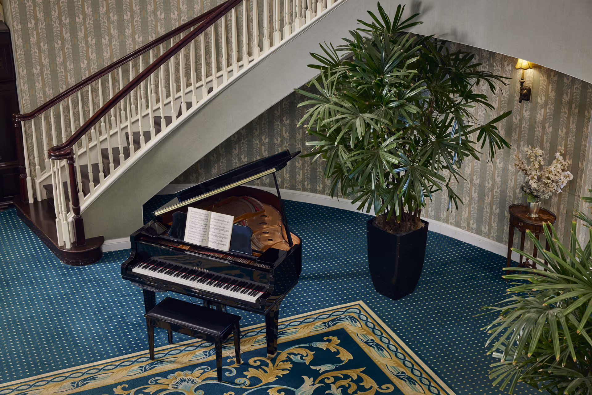 Interior view of a senior living facility featuring a black grand piano with an open sheet of music on it, a matching piano bench, a large potted plant, a small round table with a flower arrangement, and a staircase with white railings and dark wooden handrails. The floor is covered with a blue patterned carpet and a decorative rug.