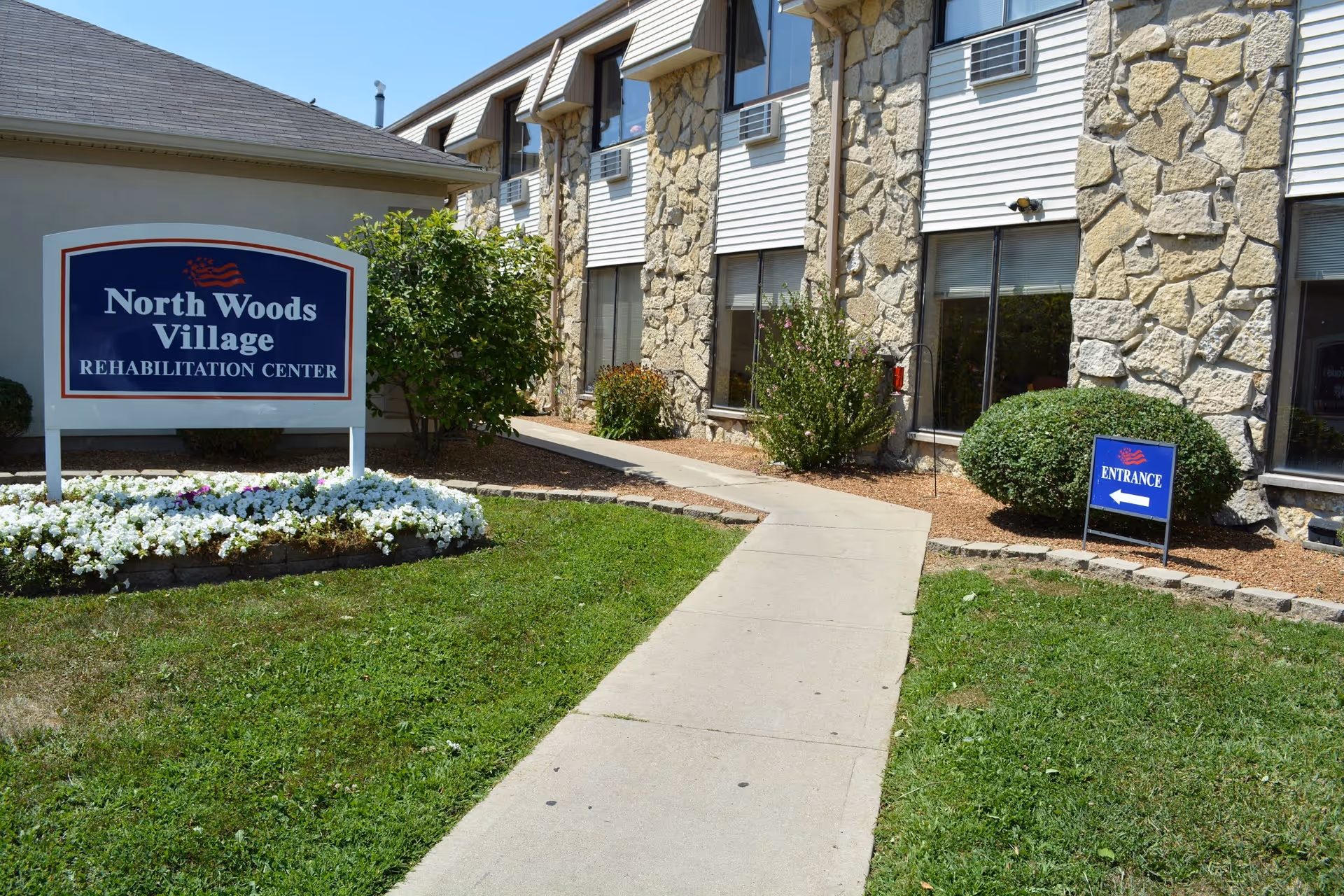 Exterior view of North Woods Village Rehabilitation Center showing a stone and siding building with windows, a concrete walkway leading to the entrance, green grass, bushes, and flower beds. There is a large sign with the facility name and a smaller sign indicating the entrance direction.