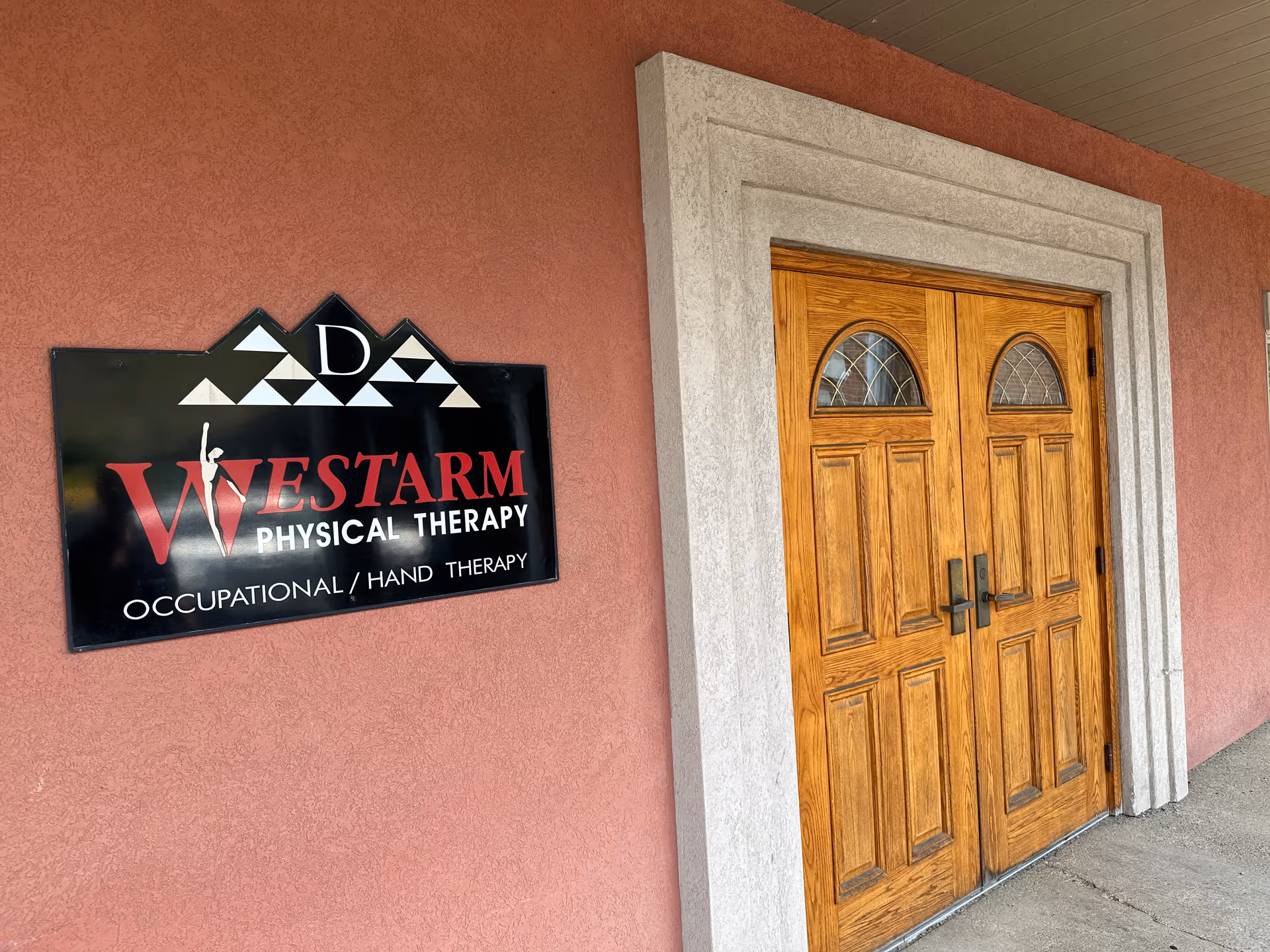 A brick exterior wall with a wooden double door and a Westarm Physical Therapy sign.