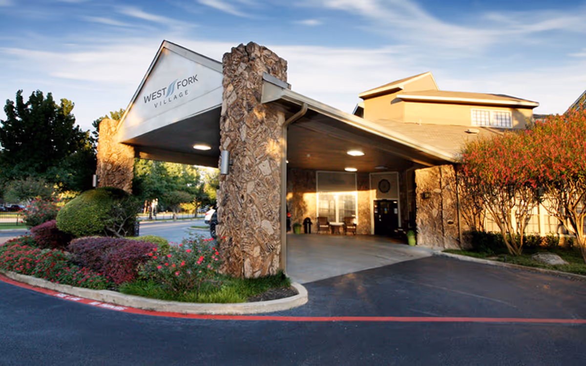 Entrance to West Fork Village senior living facility with a covered drop-off area supported by stone pillars, surrounded by landscaped bushes and trees under a partly cloudy sky.