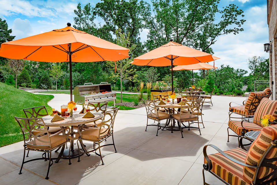 Outdoor patio with dining tables, cushioned chairs, orange umbrellas, and a grill.